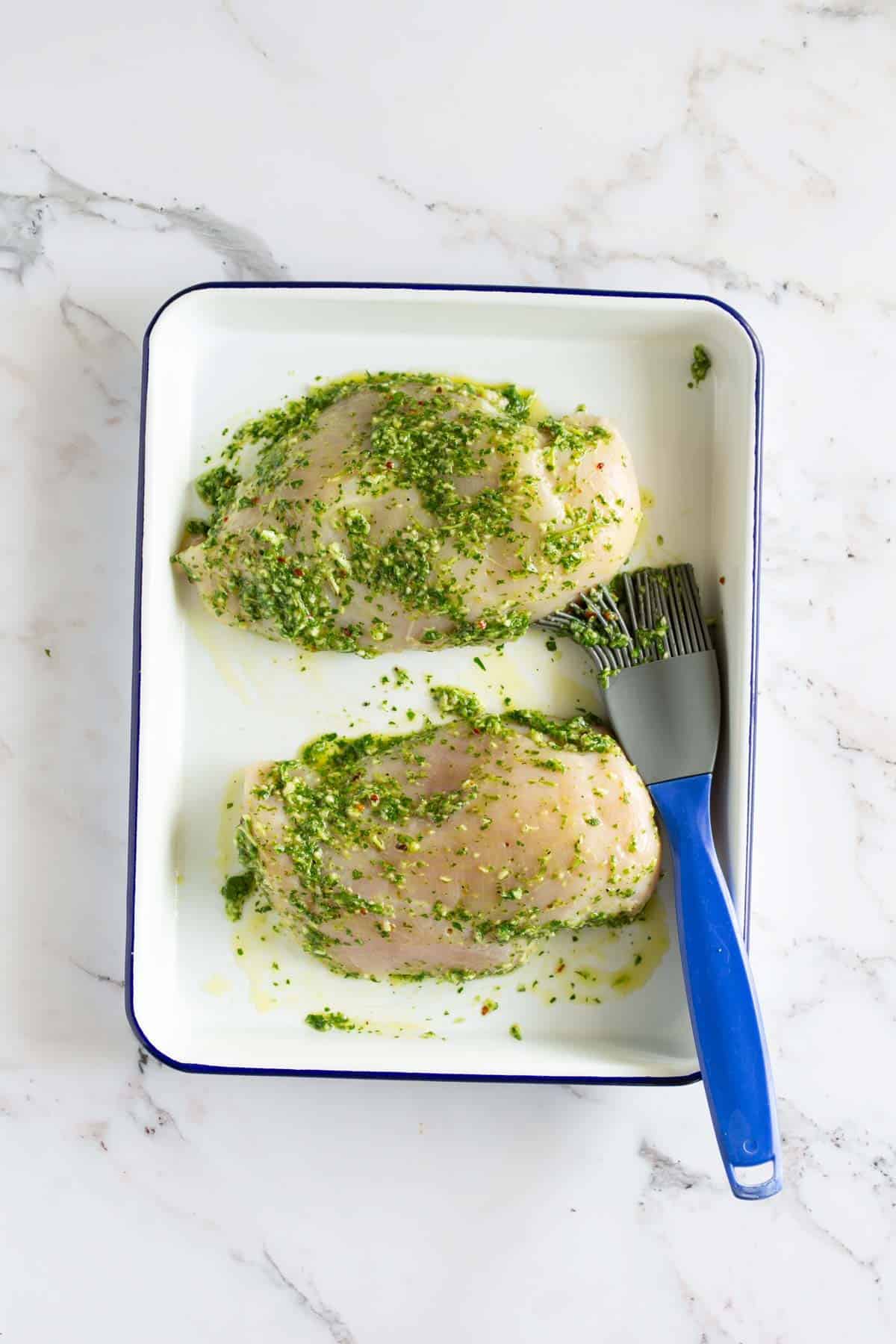Two raw chicken breasts marinated with green herbs and oil are in a white baking dish. A blue basting brush lies next to the chicken.