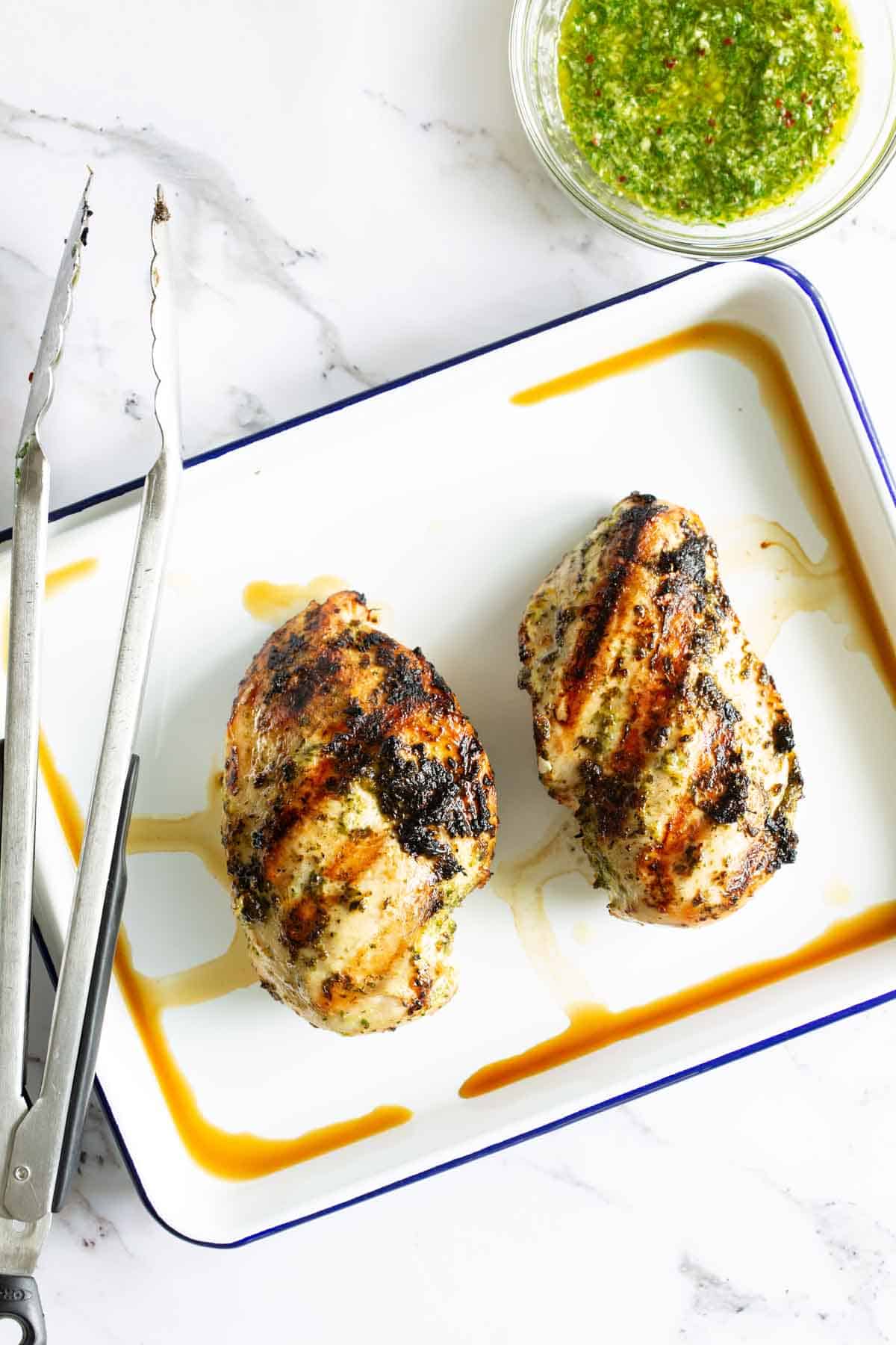 Two grilled chicken breasts on a white rectangular plate with tongs. A bowl of green sauce is placed nearby.
