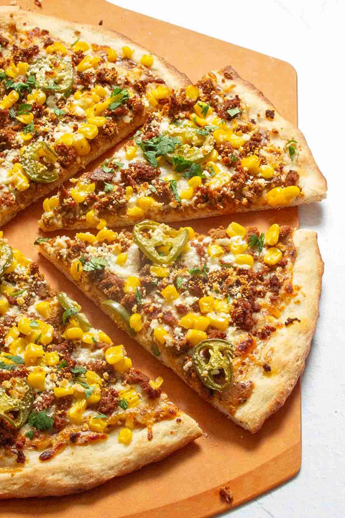 A pizza topped with corn kernels, sliced jalapenos, crumbled meat, cilantro, and cheese, sliced into triangles on a wooden board.