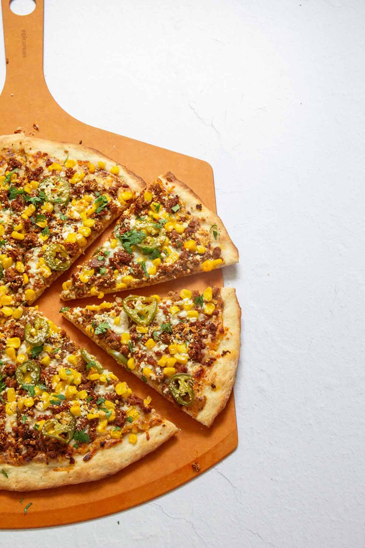 A pizza with sliced jalapeños, corn, green peppers, and ground meat on a wooden board. One piece has been partially removed.