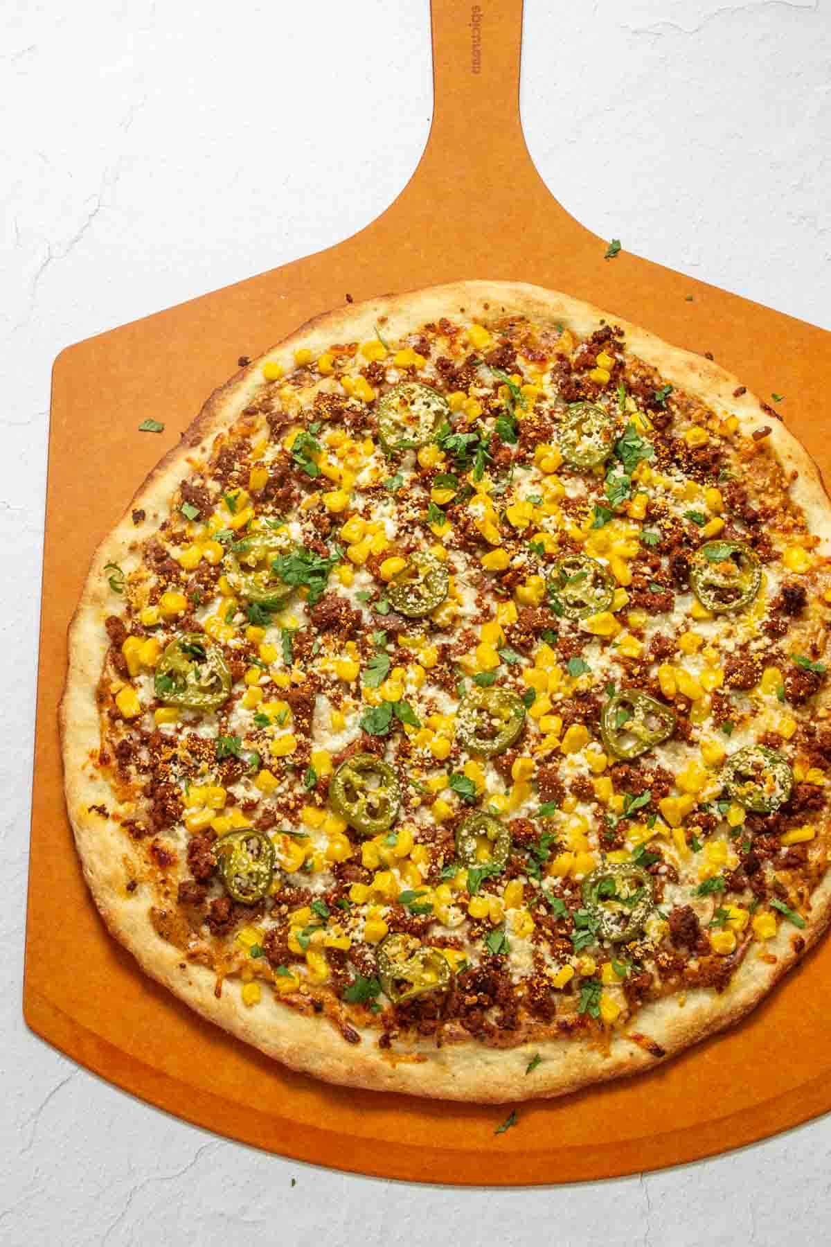 A pizza topped with corn, jalapeños, ground meat, cheese, and herbs is placed on an orange pizza peel.