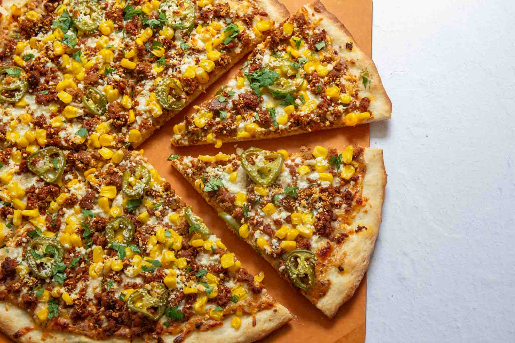 A pizza topped with ground meat, corn, jalapenos, and cheese is cut into slices on an orange surface.