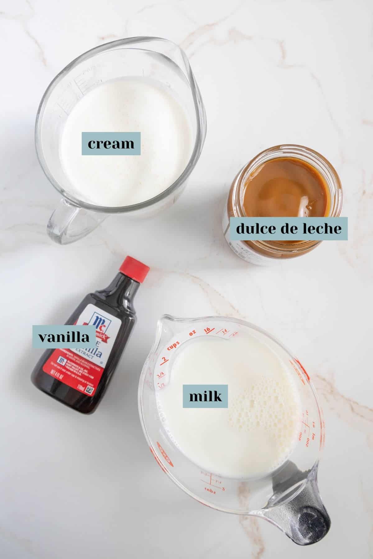 Overhead view of ingredients on a countertop: a jar of dulce de leche, a bottle of vanilla extract, a measuring cup of cream, and a measuring cup of milk.