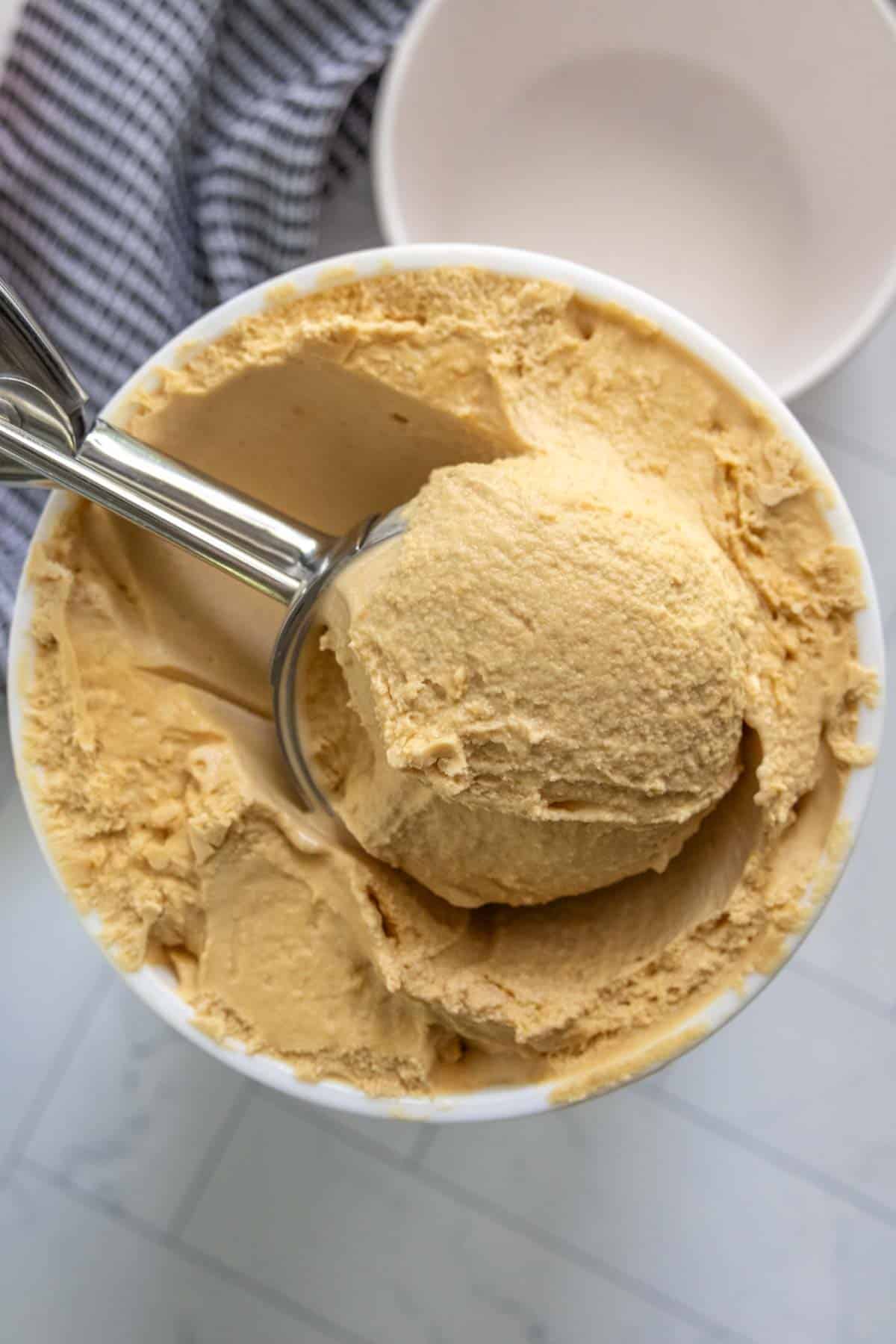 A metal scoop is inserted into a container of creamy, light brown ice cream. A white bowl and a striped cloth are partially visible in the background.