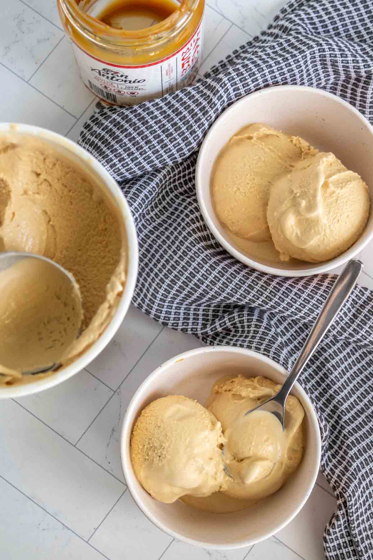 Two bowls of caramel ice cream with spoons placed on a checkered cloth, next to an open jar of caramel sauce on a tiled surface.