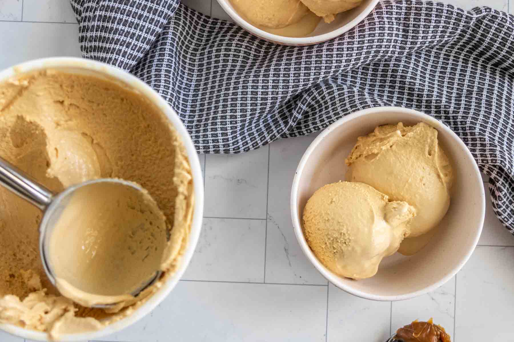 Two white bowls filled with scoops of light brown ice cream, next to a larger container of the same ice cream with a metal scooper inside, all placed on a white tiled surface with a gray cloth.