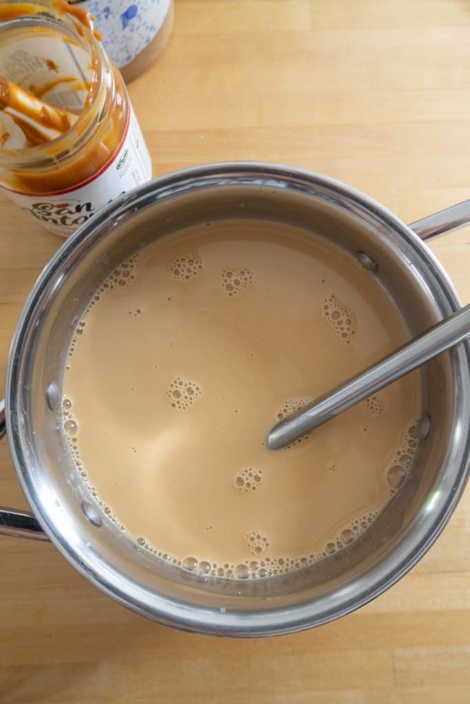 A pot filled with a light brown liquid and a metal spoon sits on a wooden surface next to an open jar containing a caramel-like spread.