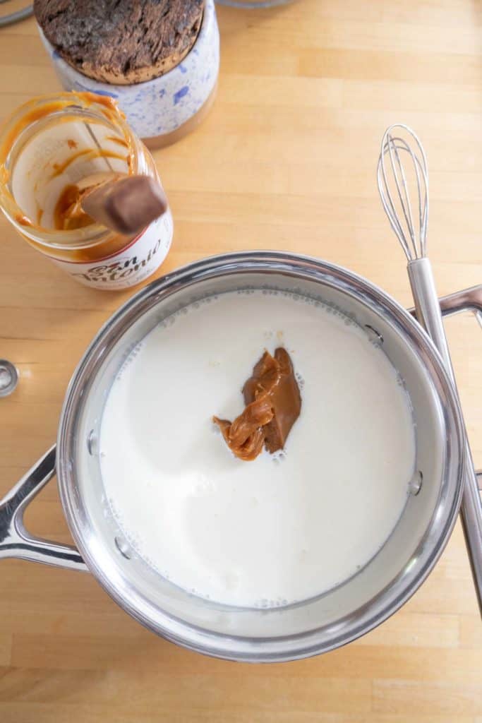 A saucepan containing milk and a dollop of caramel sits on a wooden surface next to a whisk and an open jar of caramel with some residue on it.