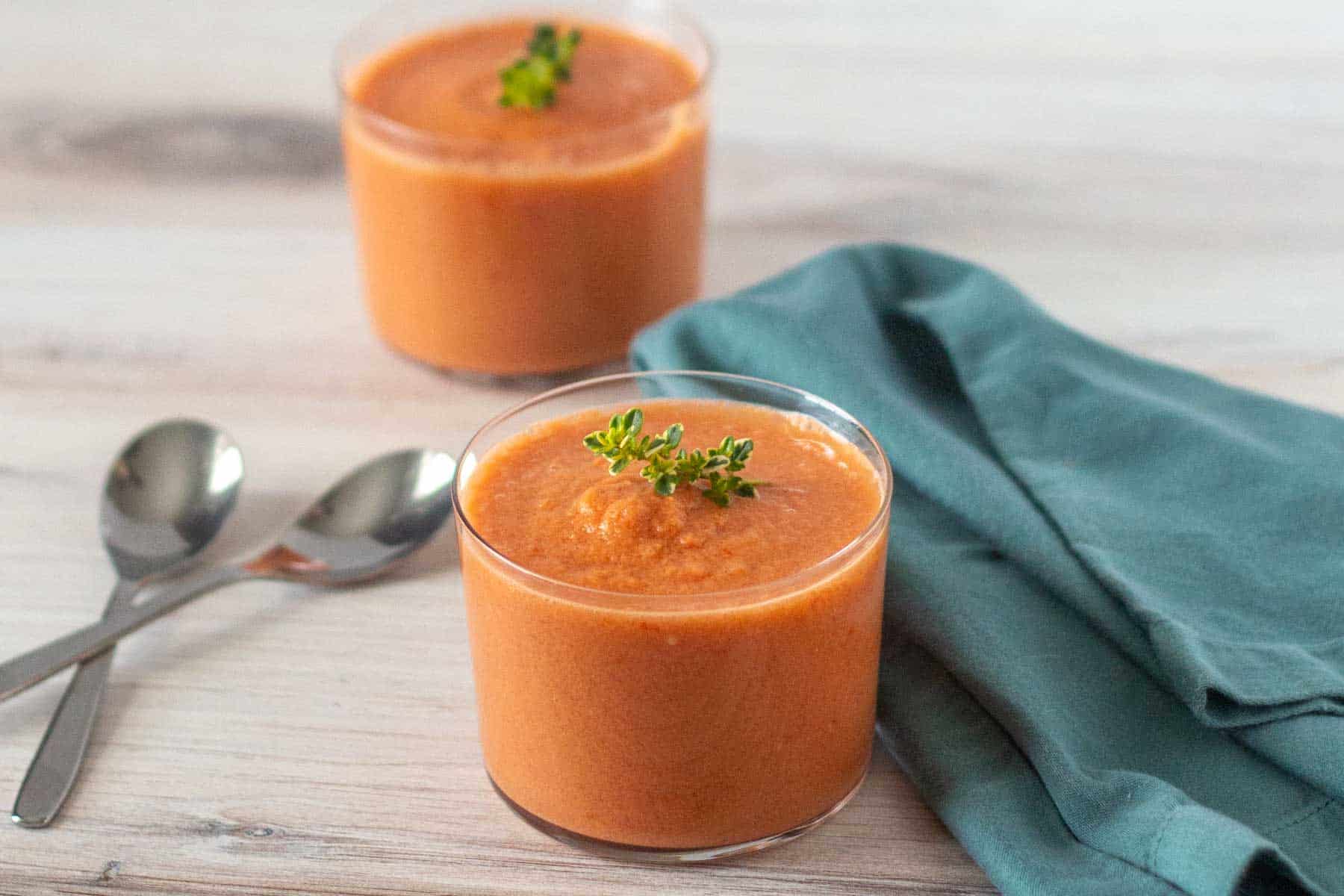 Two glasses of orange smoothie topped with green herbs, placed on a wooden surface with two spoons and a folded teal cloth nearby.