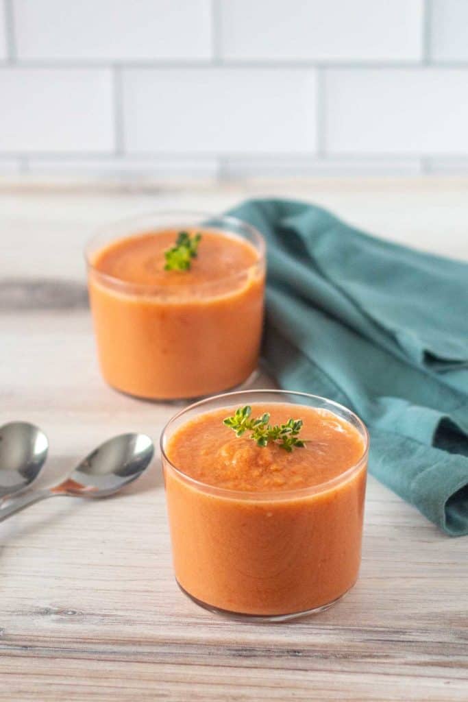 Two glass cups filled with orange-colored soup garnished with greens on a wooden surface. Two spoons and a green cloth are placed near the cups. White tile background.