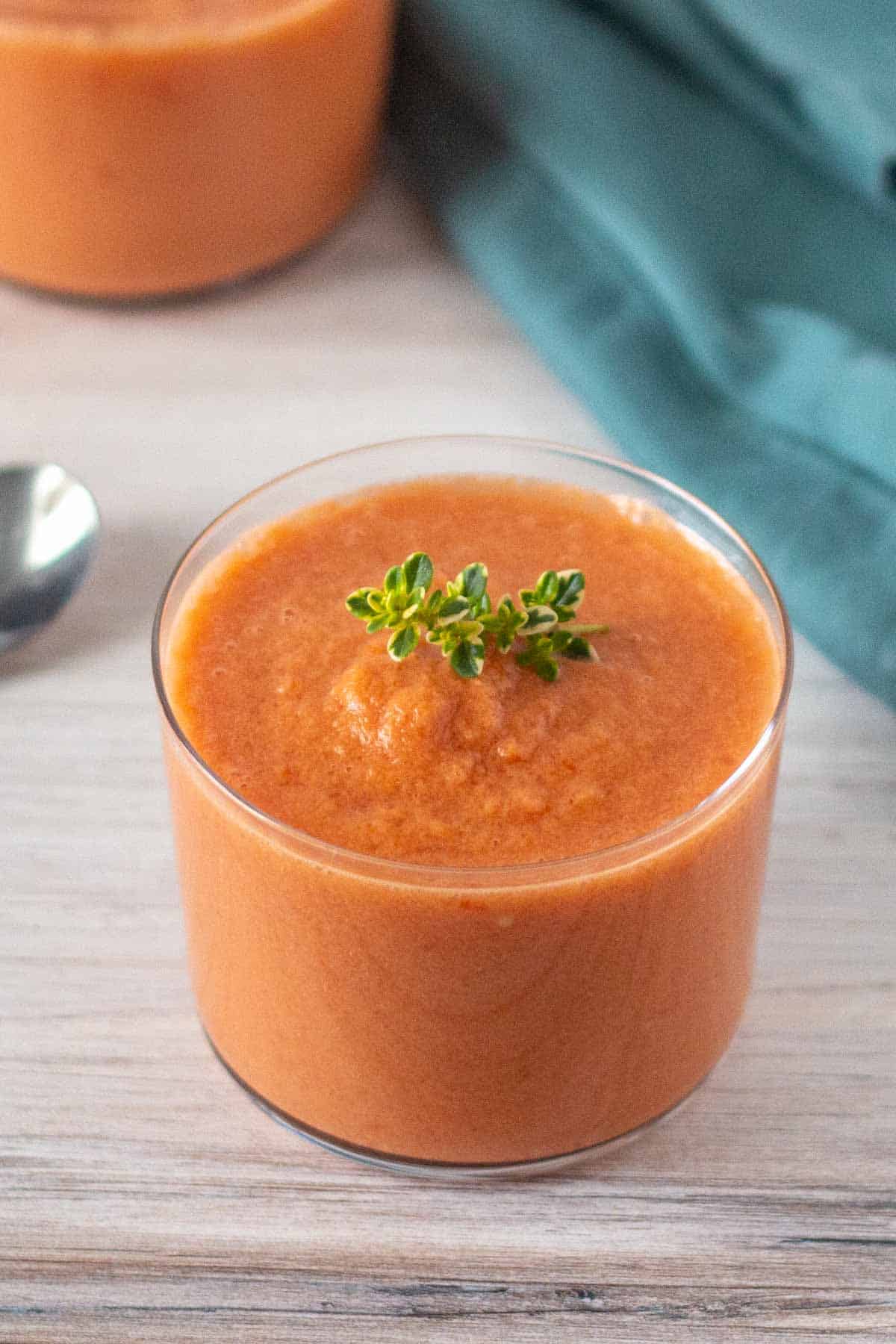 A glass filled with creamy orange soup garnished with a sprig of fresh green herbs, placed on a light wooden surface with a spoon and green cloth nearby.