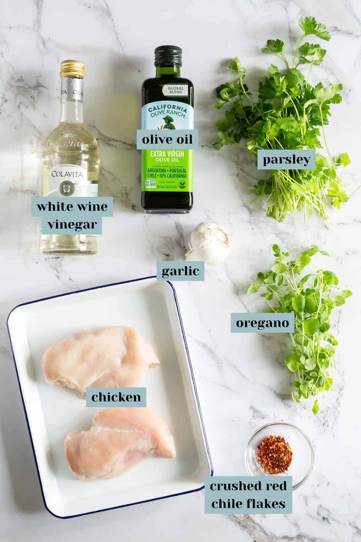 Ingredients for a dish placed on a marble surface, including white wine vinegar, olive oil, parsley, garlic, oregano, raw chicken breast, and crushed red chile flakes.