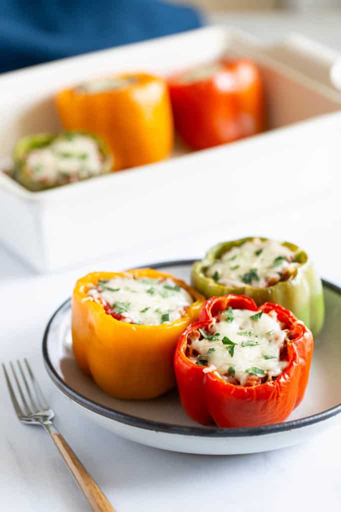 A plate of stuffed bell peppers with melted cheese, garnished with herbs, next to a baking dish with more peppers.