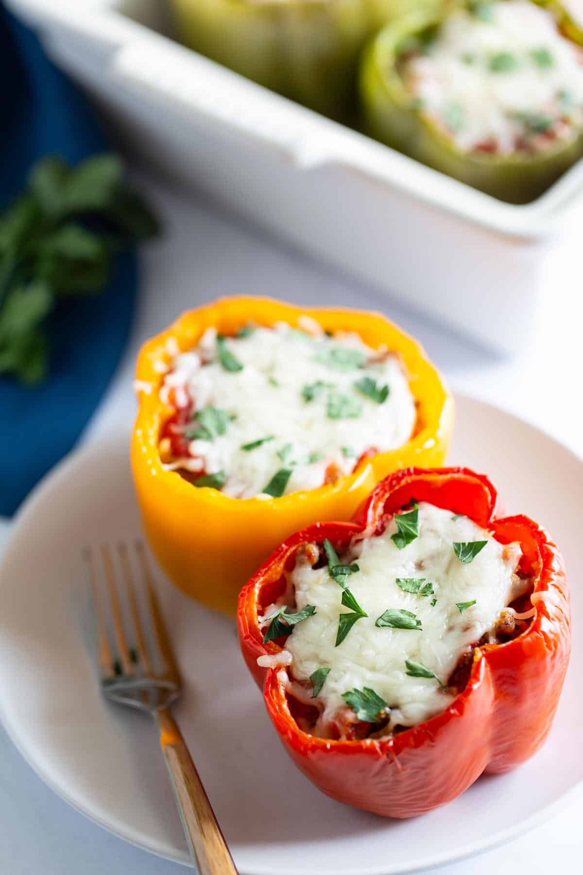 Two stuffed bell peppers on a white plate, one yellow and one red, filled with rice, meat, and herbs, topped with melted cheese.