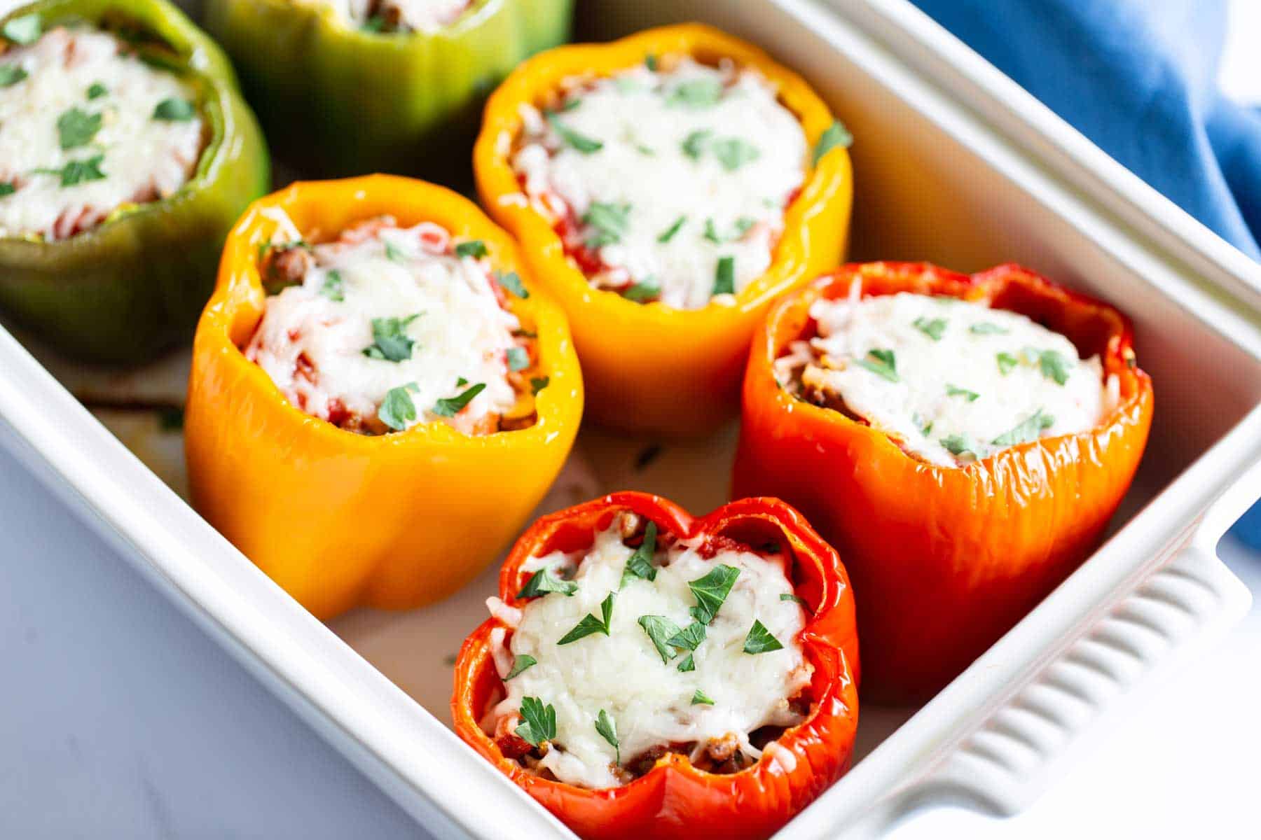 Stuffed bell peppers in red, yellow, and green, filled with rice and meat, topped with melted cheese and parsley, in a baking dish.