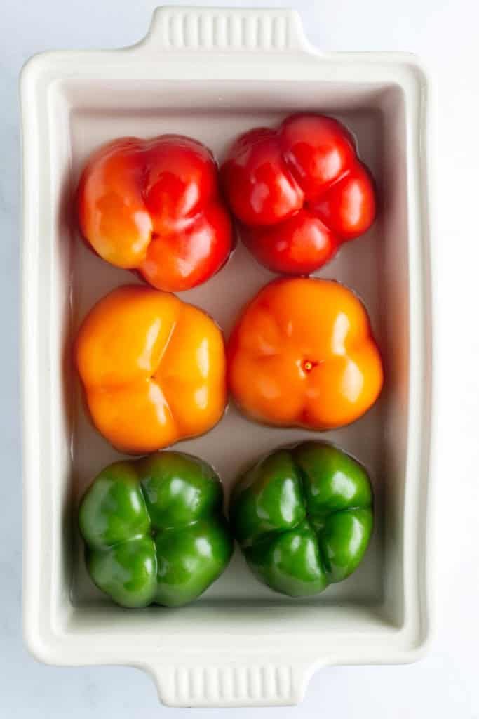 Four bell peppers, two red and two green, arranged in a white ceramic dish on a light background.