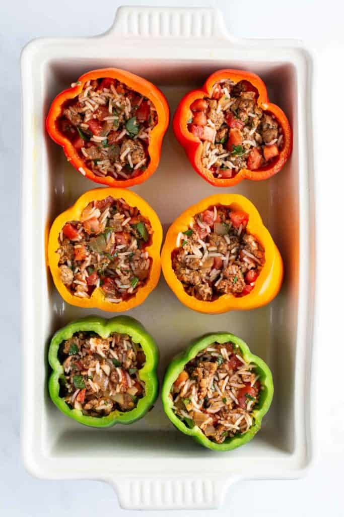 Six stuffed bell peppers with rice and vegetables in a baking dish, arranged in two rows with red, yellow, and green peppers.