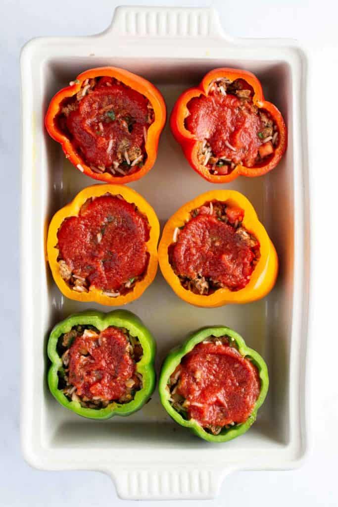 Six stuffed bell peppers in a baking dish, three red and three green, topped with tomato sauce.
