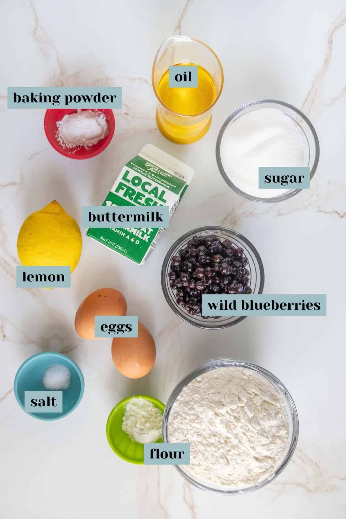 Ingredients for a recipe including baking powder, oil, sugar, buttermilk, wild blueberries, flour, eggs, a lemon, and salt, arranged on a marble surface.