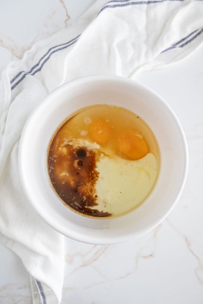 A white bowl contains a mixture of eggs, oil, and vanilla extract on a white surface with a white cloth in the background.