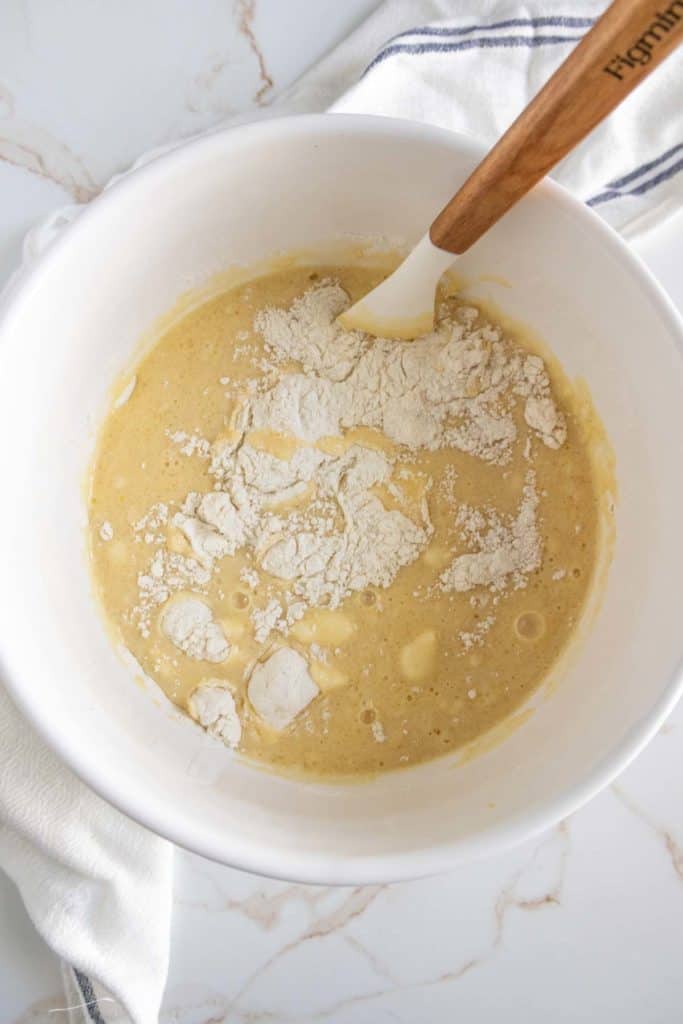 A mixing bowl with a thick yellow batter and unmixed flour on top, being stirred with a white spatula that has a wooden handle.