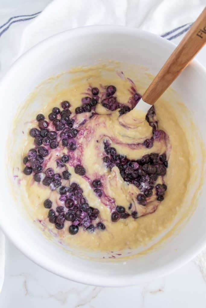 A mixing bowl containing batter with blueberries being stirred with a white and wooden spatula.