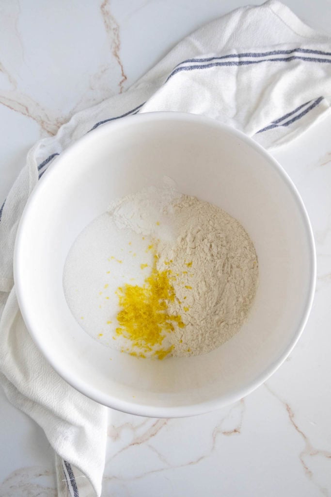 A mixing bowl with flour, sugar, and lemon zest on a white marble surface with a white cloth underneath.