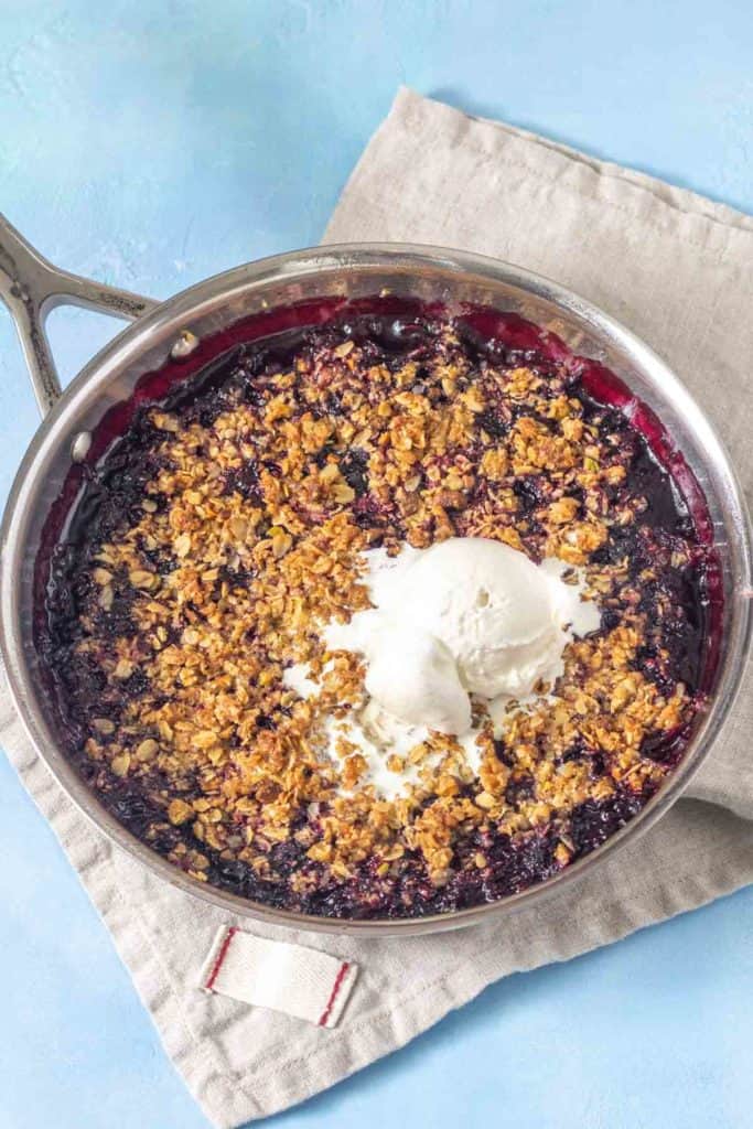A skillet containing a blueberry crumble topped with a scoop of vanilla ice cream, resting on a folded beige cloth.