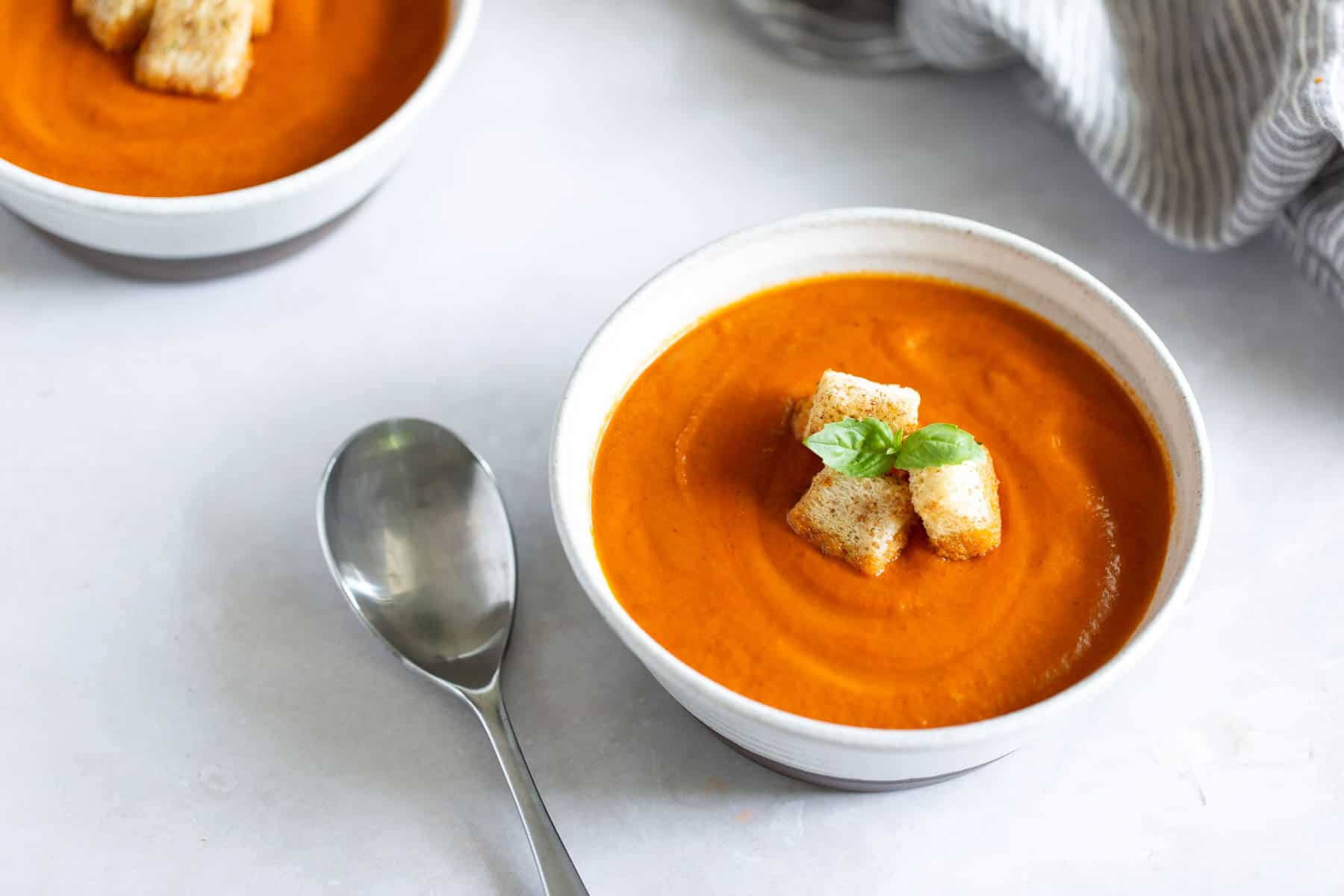 A bowl of creamy tomato soup garnished with croutons and a basil leaf, with a metal spoon placed nearby on a light surface. Another bowl of soup is partially visible in the background.