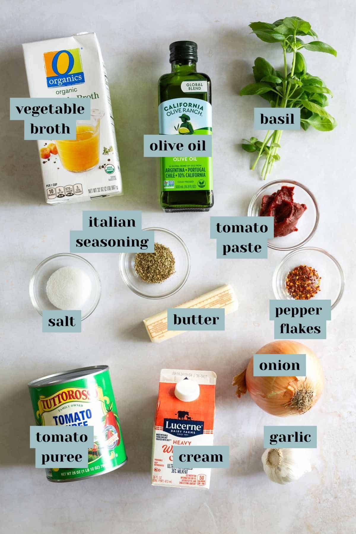 Ingredients laid out on a surface include basil, olive oil, vegetable broth, Italian seasoning, tomato paste, salt, butter, pepper flakes, onion, garlic, heavy cream, and tomato puree.