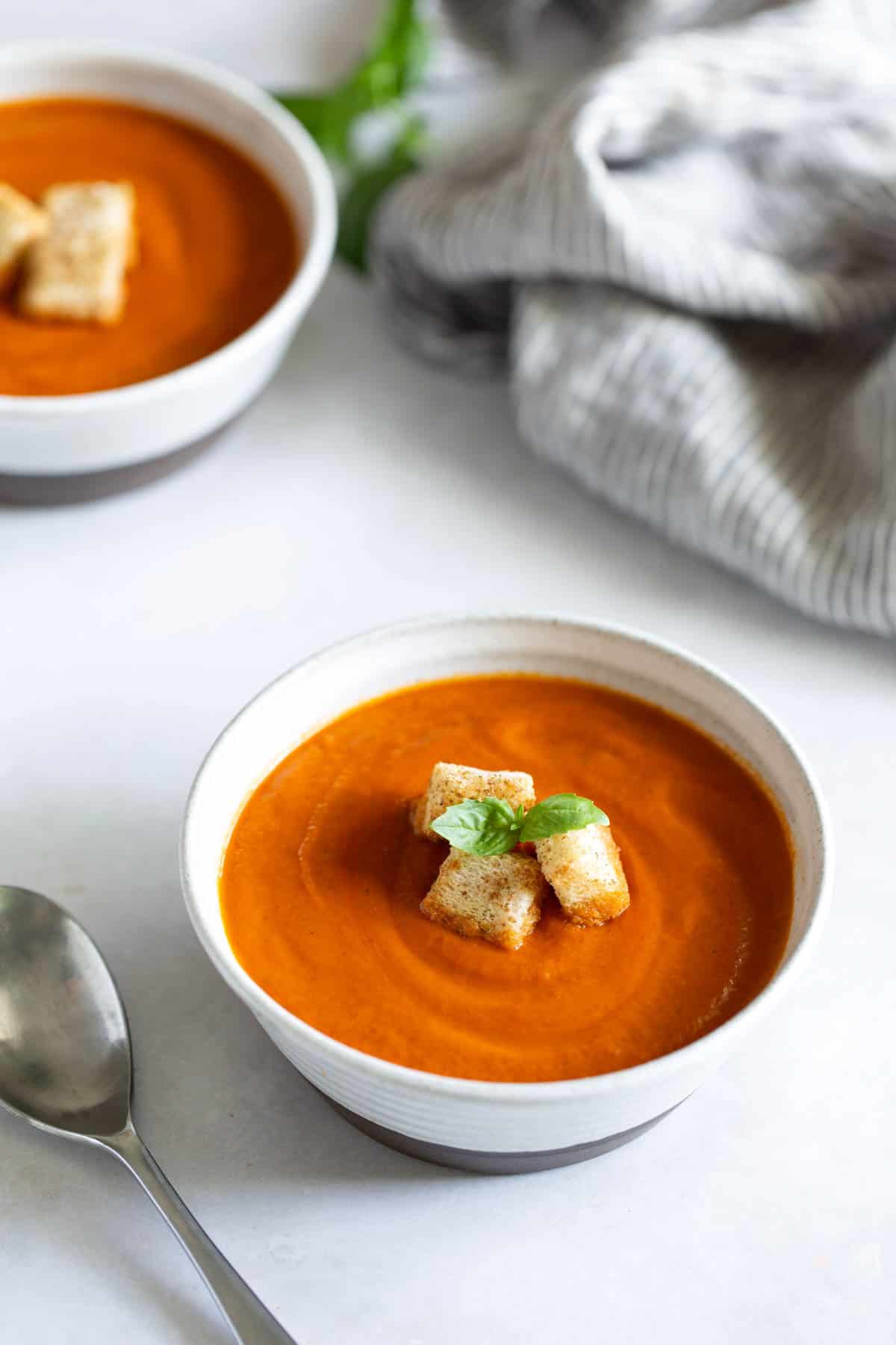 A bowl of tomato soup topped with croutons and a basil leaf, with a spoon and a striped napkin nearby. Another bowl of soup is in the background.