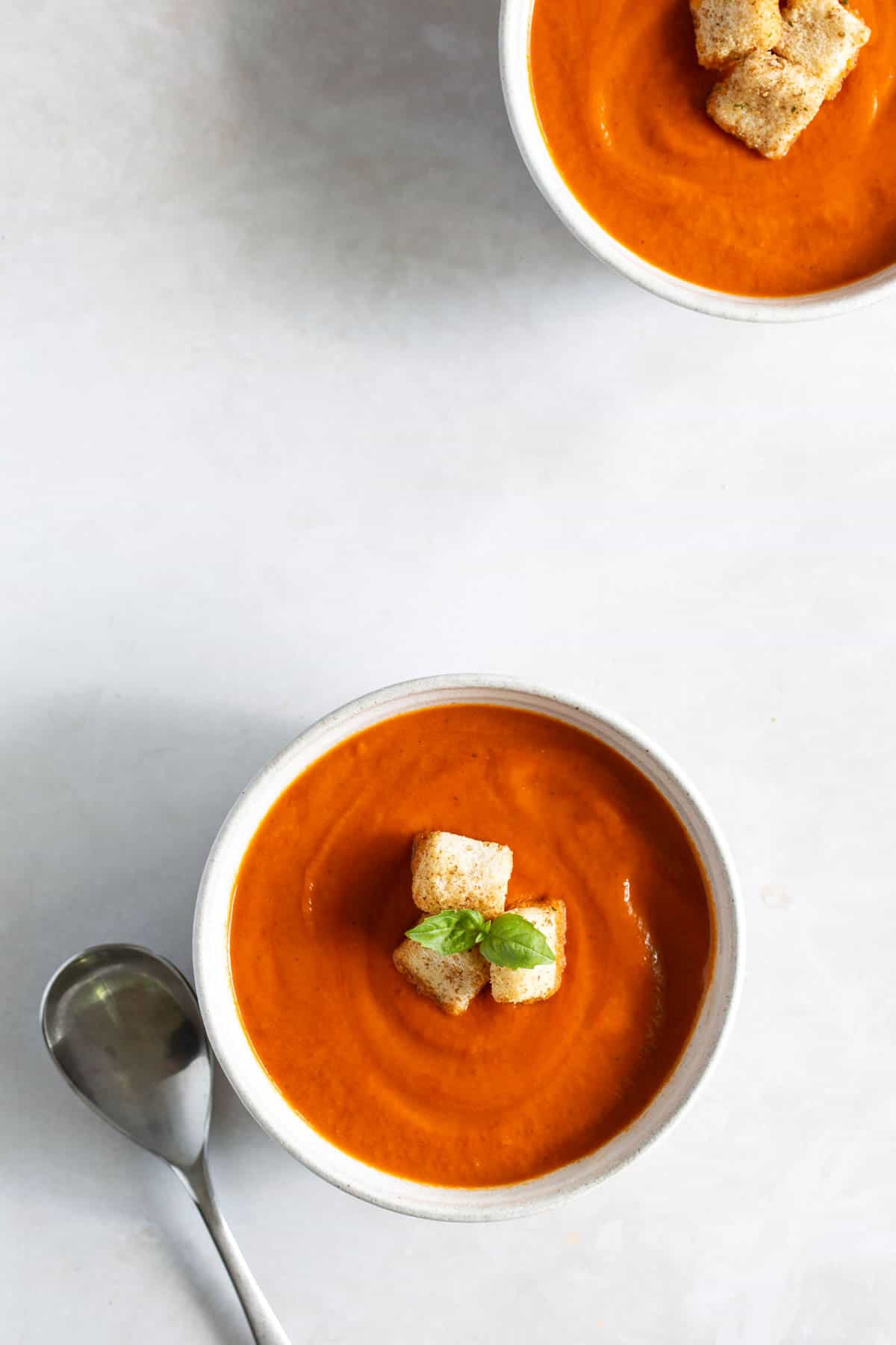 Two bowls of tomato soup with croutons and basil garnish, accompanied by a spoon on a light-colored surface.
