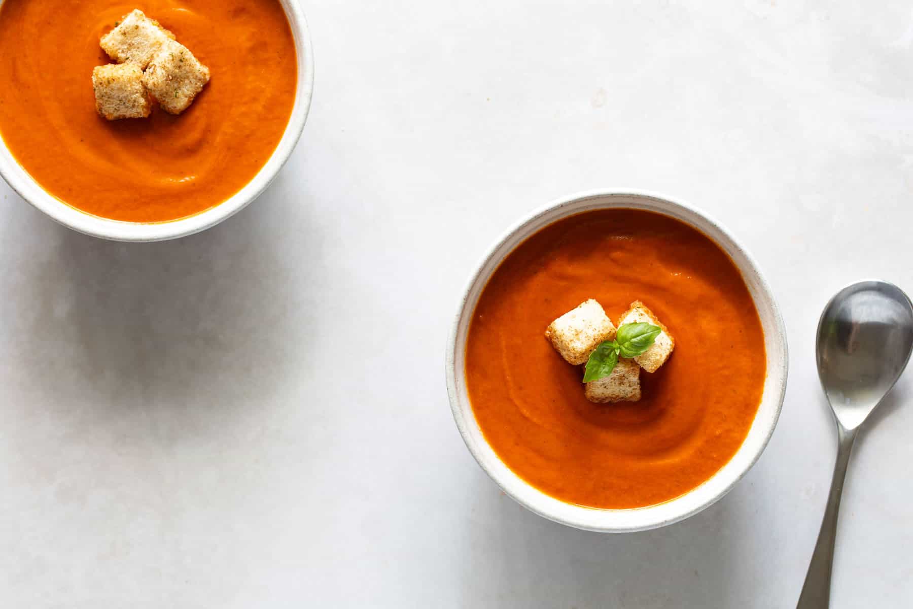 Two bowls of tomato soup topped with croutons and basil leaves, placed on a light-colored surface with a spoon beside one of the bowls.