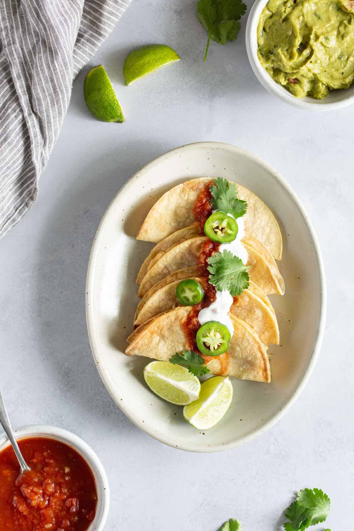 A plate of tacos topped with salsa, sour cream, and sliced jalapeños, garnished with lime wedges. Guacamole, a bowl of salsa, lime wedges, and a striped cloth are placed around the plate.