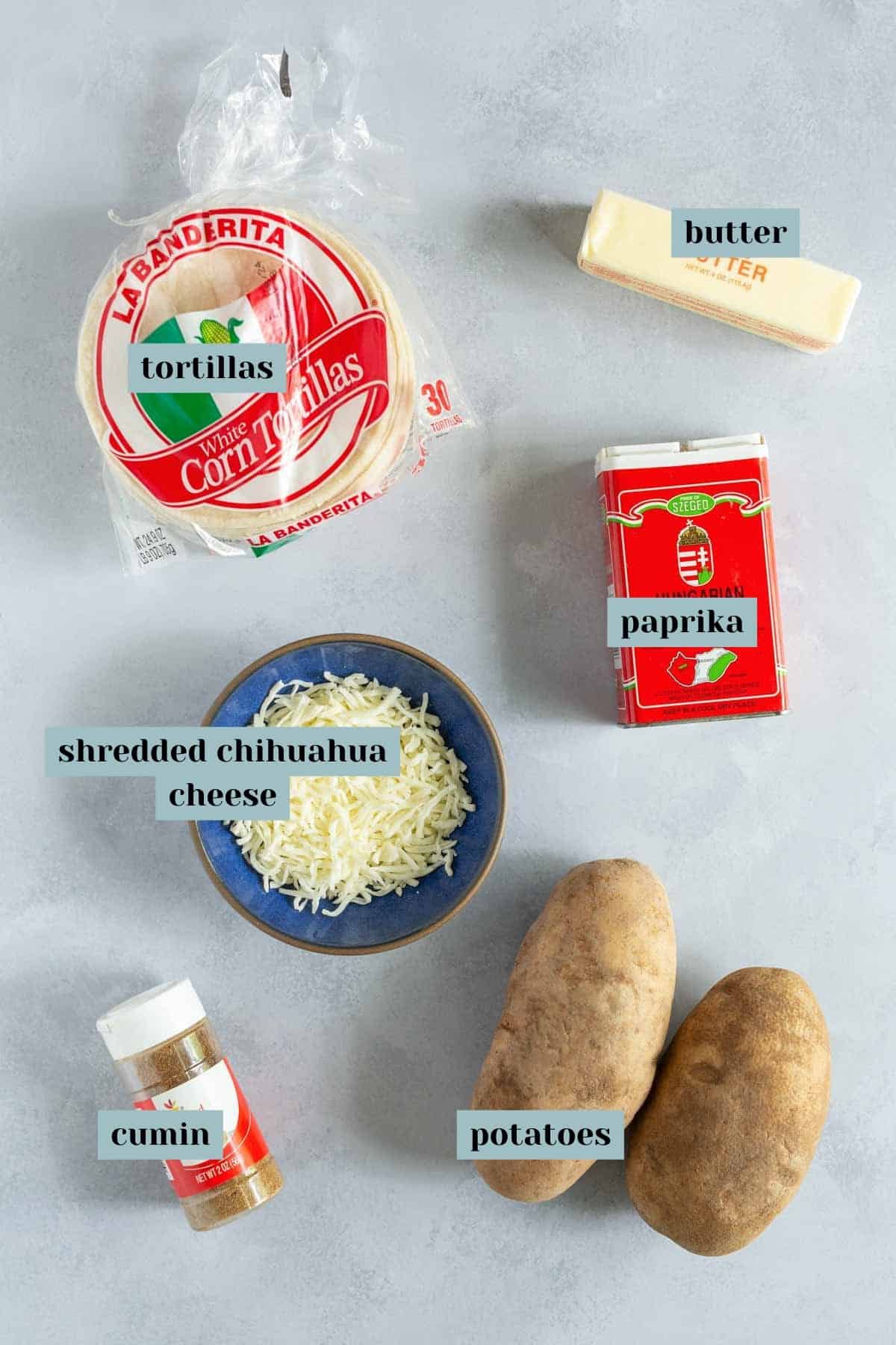 Ingredients laid out on a gray surface: a stack of tortillas in packaging, a stick of butter, a can of paprika, two russet potatoes, a bowl of shredded Chihuahua cheese, and a jar of cumin.
