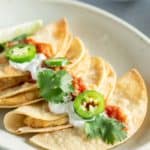 A plate of tacos topped with sliced jalapeños, cilantro, salsa, and a dollop of cream, with additional salsa in a small bowl in the background.