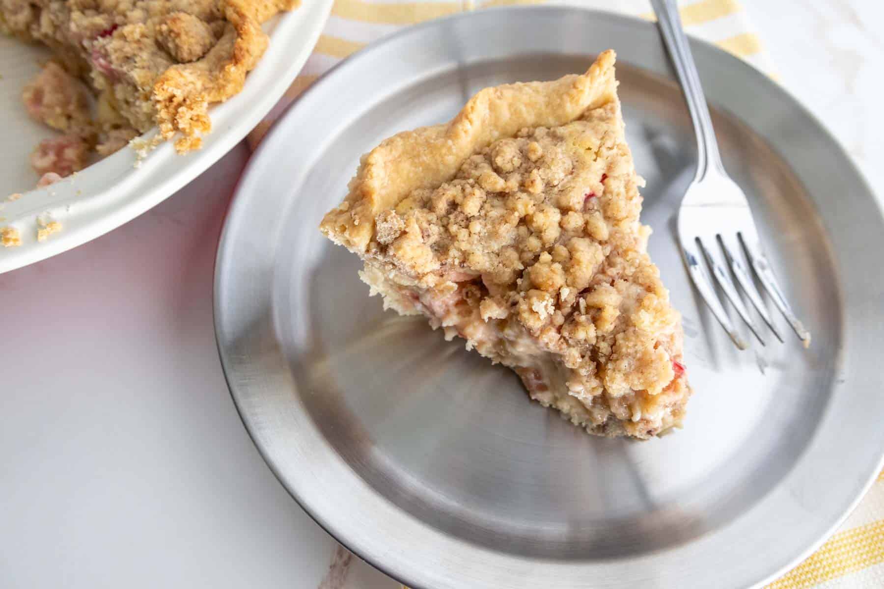 A slice of crumb-topped pie on a silver plate with a fork beside it, positioned next to the remaining pie in a white dish.