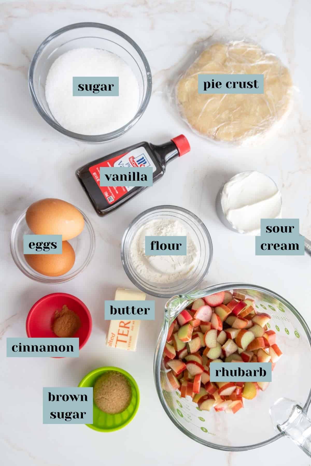 Top-down view of ingredients for a rhubarb pie, including sugar, pie crust, vanilla, eggs, flour, sour cream, cinnamon, butter, brown sugar, and chopped rhubarb in a measuring cup.