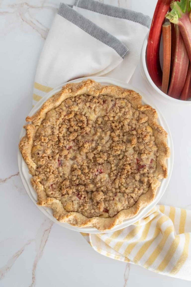 A baked crumb-topped pie on a white surface with a striped cloth underneath, next to a bowl containing stalks of fresh rhubarb.