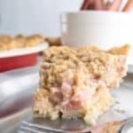 A slice of crumb-topped pie with a creamy and chunky filling sits on a silver plate with a fork in the foreground. In the background, there is a container filled with rhubarb stalks.