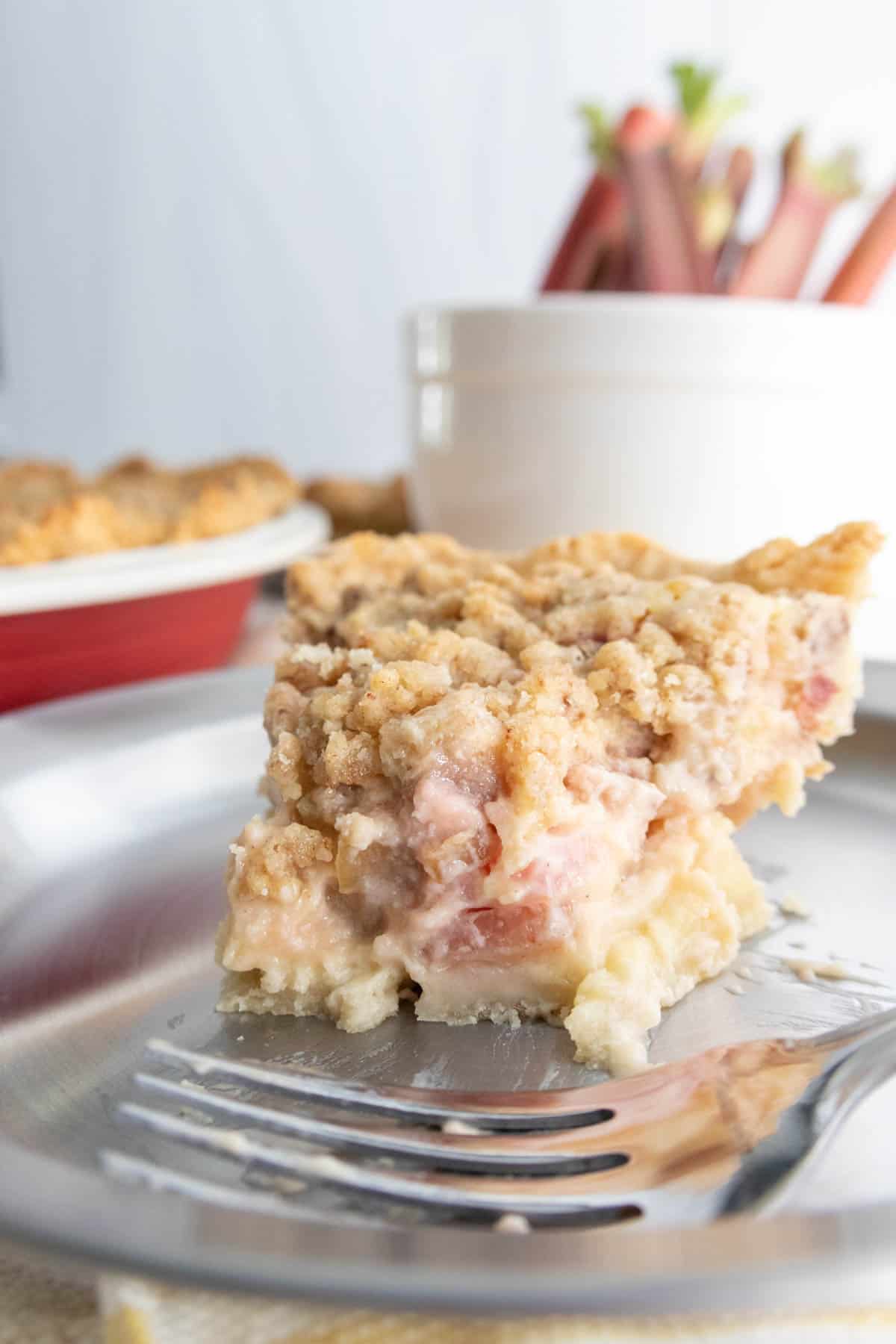 A slice of crumb-topped pie with a creamy and chunky filling sits on a silver plate with a fork in the foreground. In the background, there is a container filled with rhubarb stalks.