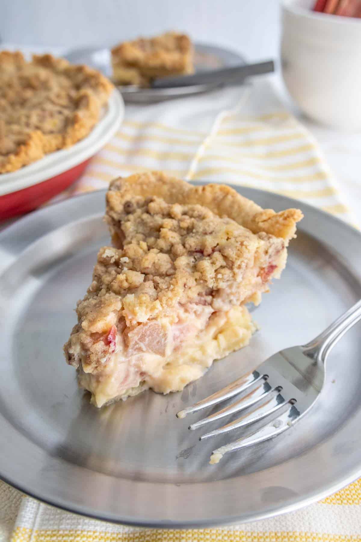 A slice of pie with crumb topping on a metal plate, with a fork resting beside it. A whole pie and another slice are in the background.