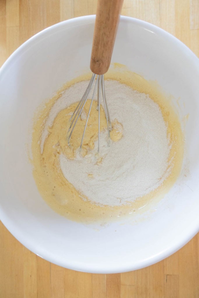 A metal whisk in a white mixing bowl with a mixture of dry and wet ingredients on a wooden surface.