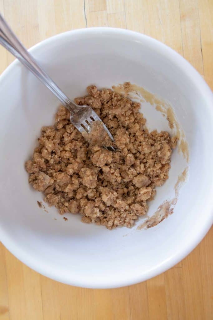 A white bowl containing crumbly brown mixture being stirred with a fork on a wooden surface.