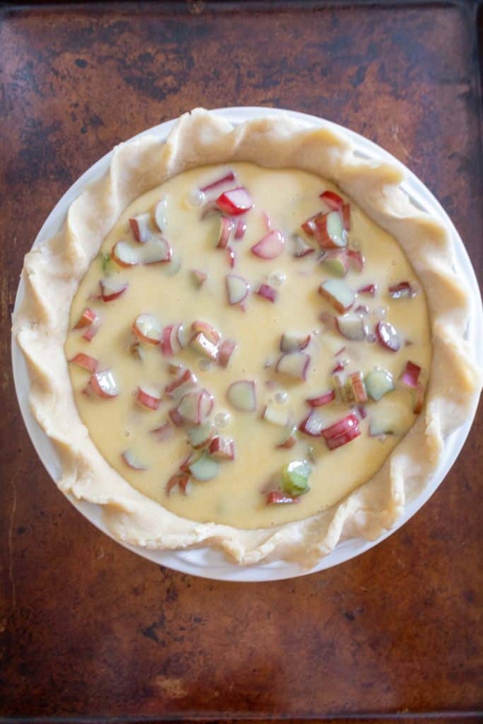 A raw pie crust filled with an egg mixture and sliced vegetables, ready to be baked. The pie is placed on a baking sheet.