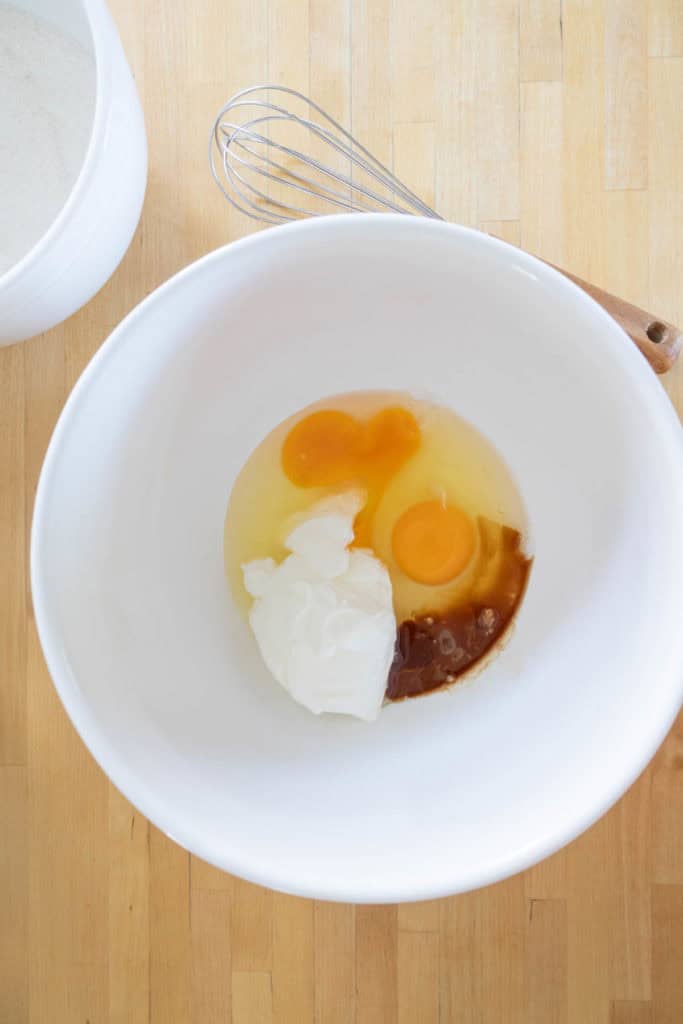 A white bowl contains two cracked eggs, vanilla, and sour cream on a wooden surface with a whisk and another bowl nearby.