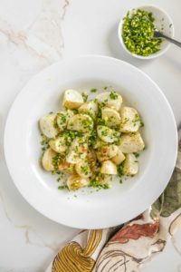 A white plate filled with roasted potatoes sprinkled with chopped herbs. A small bowl with additional herbs and a spoon are placed nearby on the marble countertop. A patterned cloth is partially visible.