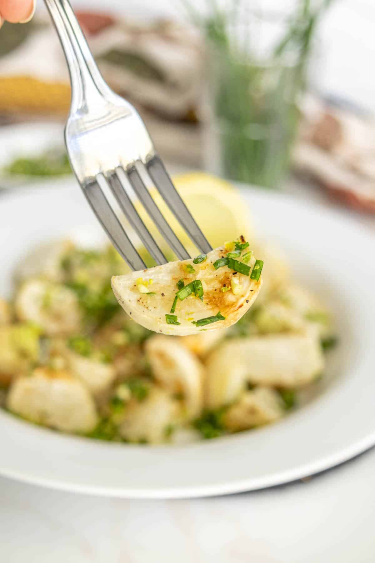 A fork holds a piece of seasoned potato gnocchi above a white plate filled with the dish. The gnocchi is garnished with chopped herbs.