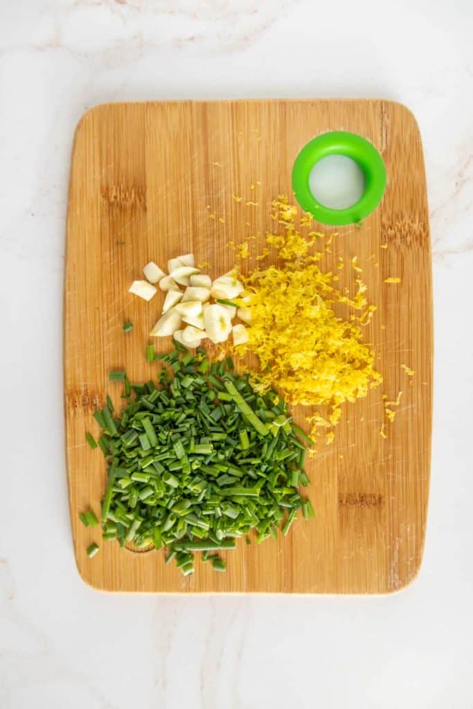 A wooden cutting board with chopped chives, minced garlic cloves, lemon zest, and a small round green container with a light-colored substance inside.
