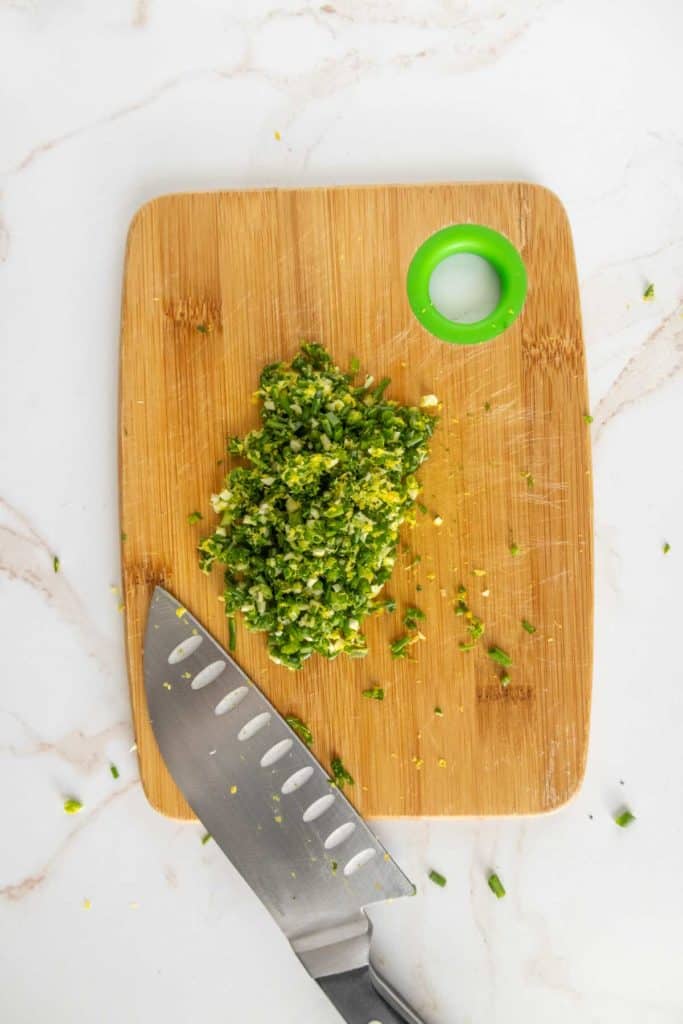 Chopped herbs on a wooden cutting board with a knife next to them on a marble surface.